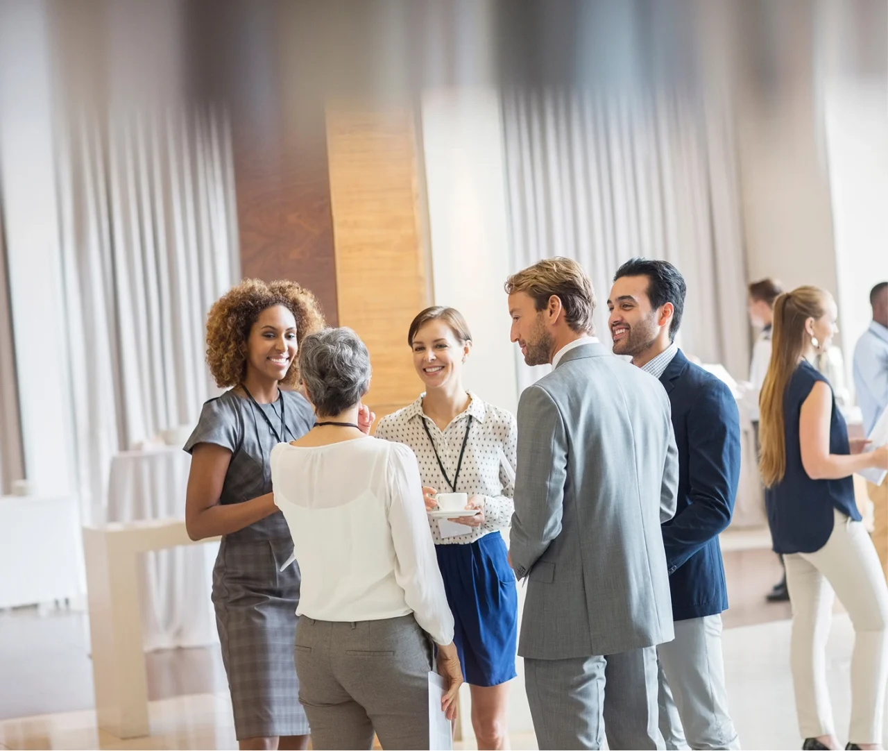 Das Bild zeigt eine professionelle Networking-Veranstaltung oder Business-Meeting in einem hellen, modernen Raum mit hohen Fenstern und bodenlangen, halbtransparenten Vorhängen. Im Vordergrund stehen fünf Personen in Business-Kleidung in einer lockeren Gesprächsrunde. Eine Frau mit lockigen Haaren in einem grauen Kleid unterhält sich mit einer Kollegin mit Kurzhaarschnitt und weißem Oberteil, die von hinten zu sehen ist. In der Mitte steht eine lächelnde Frau in gepunkteter Bluse und blauem Rock, die eine Kaffeetasse hält und ein Namensschild trägt. Rechts daneben stehen zwei Männer in hellgrauen Anzügen – einer von hinten, der andere im Profil sichtbar.