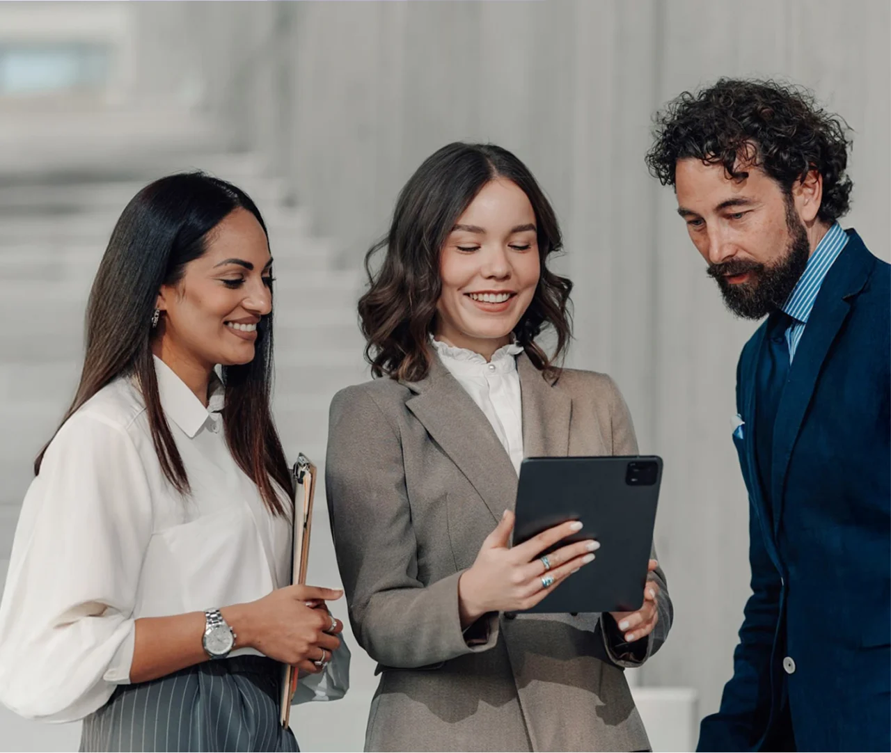 Das Bild zeigt drei Business-Professionals in formeller Kleidung, die gemeinsam auf ein Tablet schauen. In der Mitte steht eine lächelnde Frau in einem beigefarbenen Hosenanzug mit weißer Bluse, die das Tablet hält. Links neben ihr steht eine Kollegin in cremefarbener Bluse mit gestreifter Hose, die ein Clipboard in der Hand hält und eine Armbanduhr trägt. Rechts steht ein Mann mit Bart und dunklen lockigen Haaren in einem dunkelblauen Anzug mit hellblauem gestreiftem Hemd. Alle drei wirken konzentriert und interessiert an den Inhalten auf dem Bildschirm.