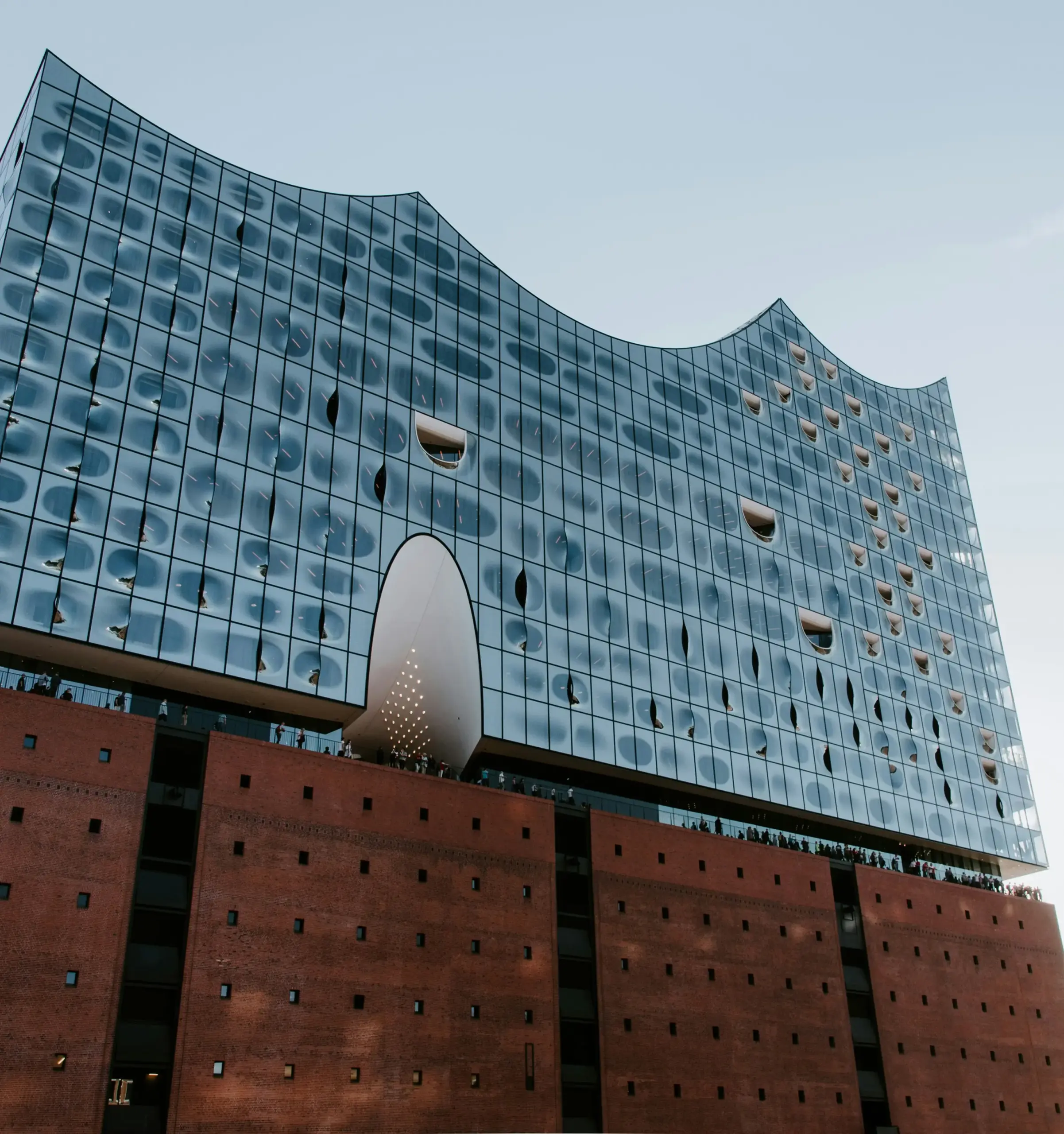 Moderne Architekturaufnahme der Elbphilharmonie in Hamburg an einem bewölkten Tag. Das ikonische Konzerthaus zeigt seine charakteristische wellenförmige Glasfassade mit reflektierenden blauen Fensterflächen, die den Himmel spiegeln.