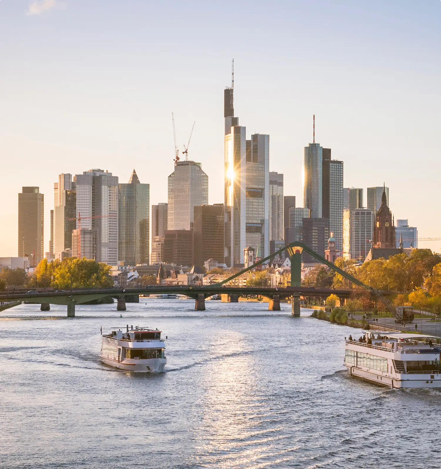 Panoramablick auf die Frankfurter Skyline mit modernen Bankentürmen bei goldenem Sonnenuntergang, Fahrgastschiffe auf dem Main und der Flößerbrücke im Vordergrund.