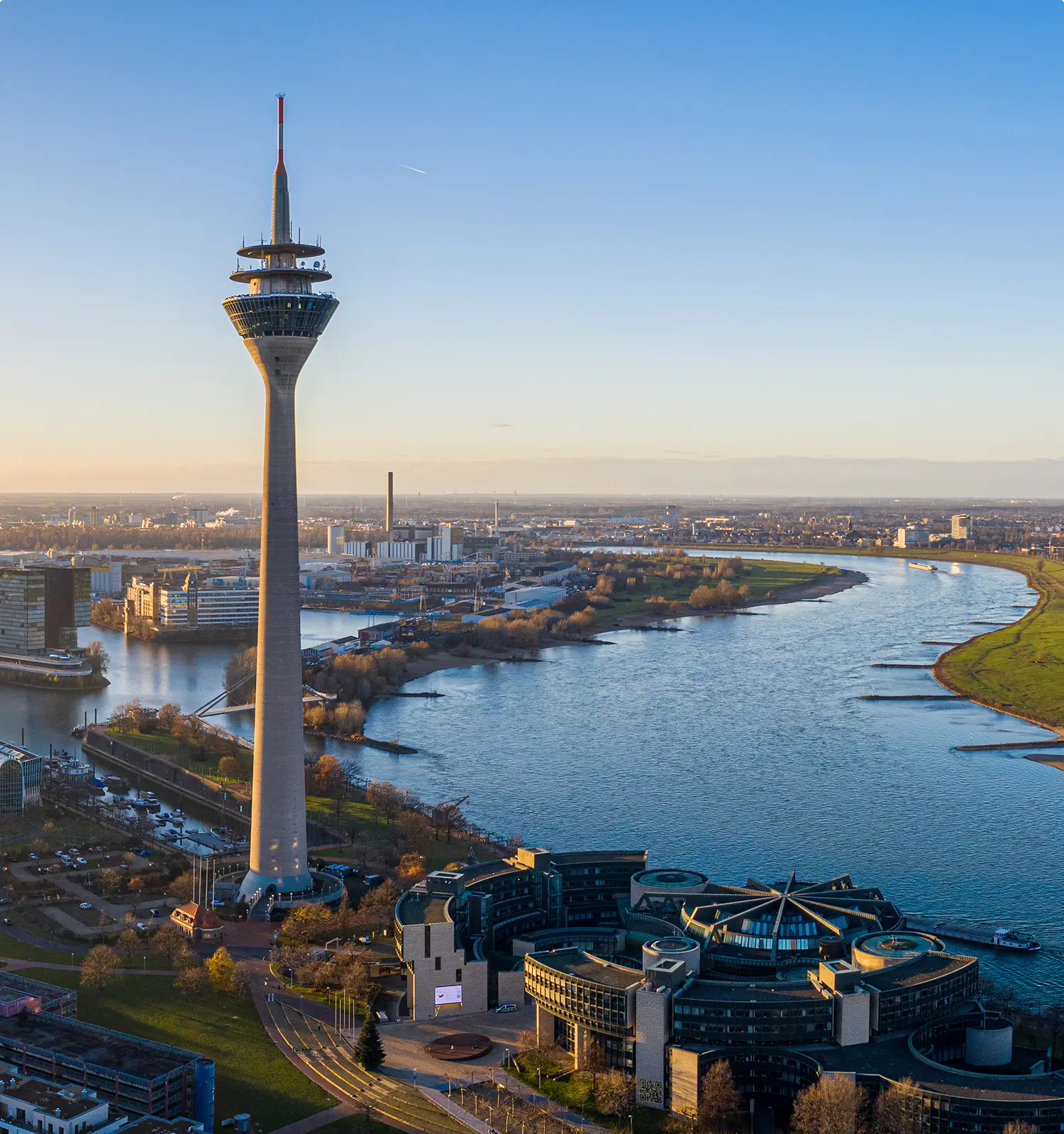 Luftaufnahme des Düsseldorfer Rheinturms und des Medienhafens mit moderner Architektur entlang des Rheins bei goldenem Sonnenuntergangslicht unter klarem Himmel.