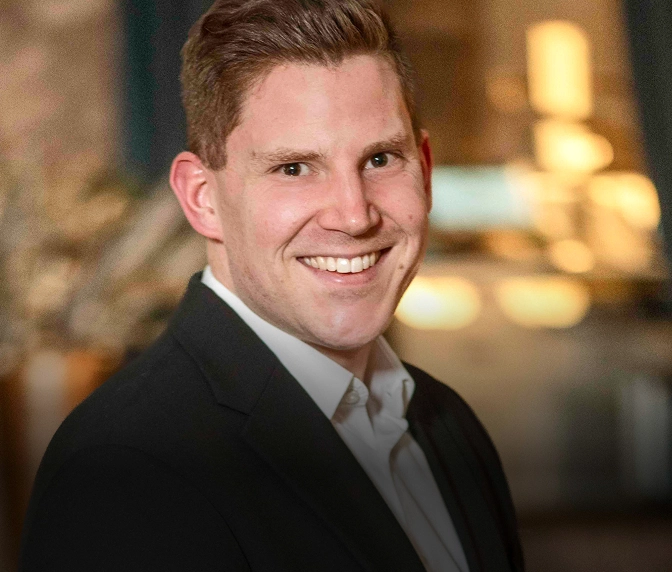 Philip Knopf - Professionelles Porträt eines lächelnden jungen Mannes in schwarzem Anzug mit weißem Hemd vor unscharfem Innenraum mit warmer Beleuchtung.